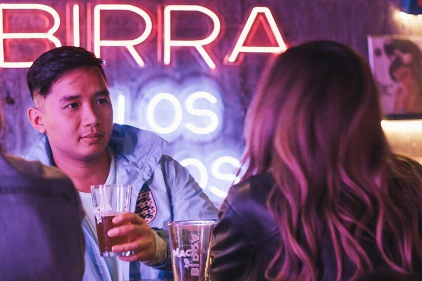 A person holding a glass with a beverage, a neon sign in the background, and another person partially visible. 1