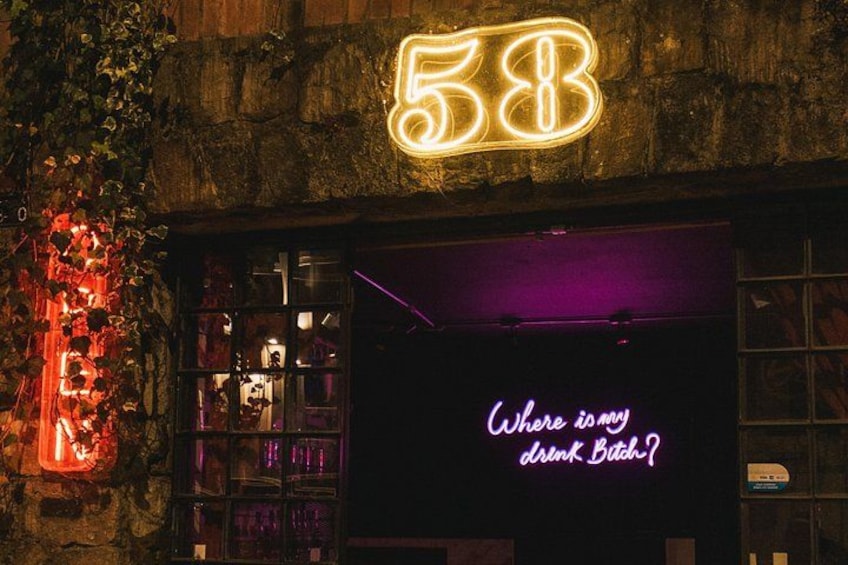 Un letrero de neón con el número 58, un letrero de neón con el texto 'Where my drink Bitch?', y una pared de ladrillo con plantas trepadoras. 3