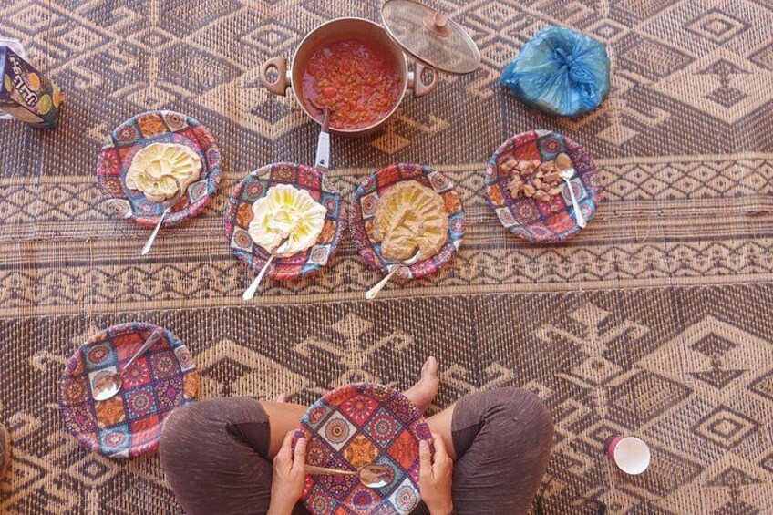 A person sitting on a patterned carpet with plates of food and a pot on the floor. 2
