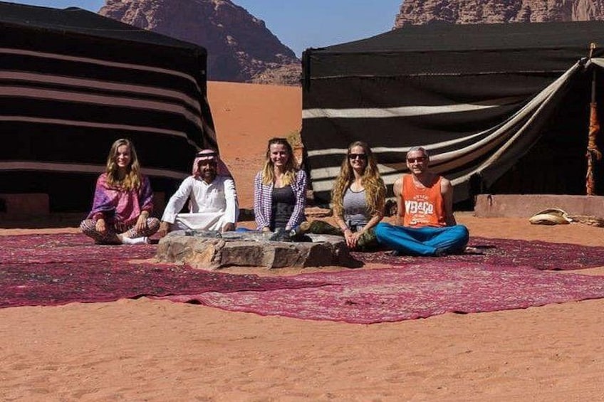 A group of people sitting on a red carpet in a desert with tents in the background. 4