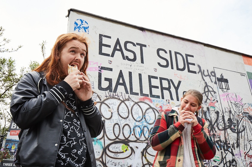Due persone che mangiano davanti a un muro ricoperto di graffiti con la scritta 'EAST SIDE GALLERY'. 1