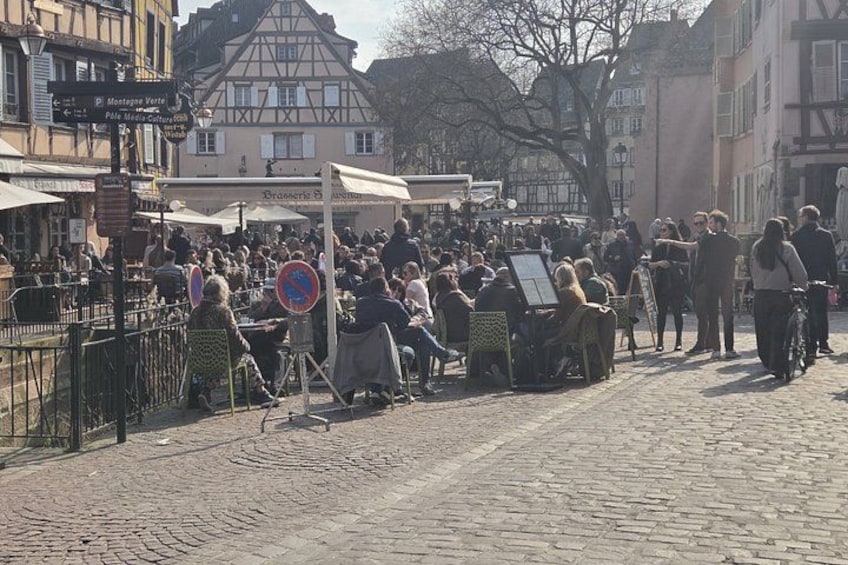 A crowded outdoor market with people sitting and standing, surrounded by buildings and cobblestone streets. 5