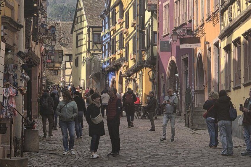 Calle adoquinada bordeada por edificios de colores, gente caminando y mesas al aire libre en cafés. 2