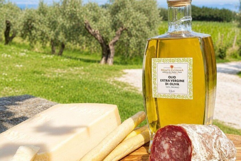 Une bouteille d’huile d’olive extra vierge Monte Cincignano, un morceau de fromage et une tranche de salami, le tout posé sur une surface en bois, avec un olivier en arrière-plan. 4