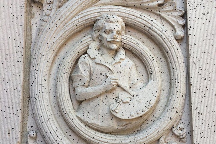 Een gedetailleerd stenen beeldhouwwerk van een figuur die een rol met tekst vasthoudt, omringd door decoratieve versieringen. 4