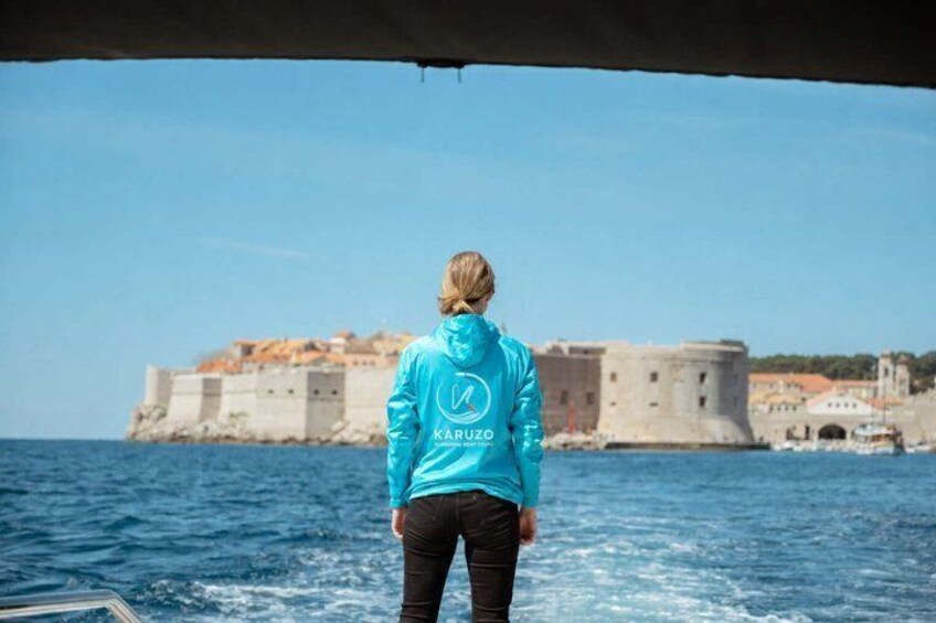 A person in a blue jacket with 'KARUZO' on the back, standing on a boat with a view of a historic fort and the sea. 5