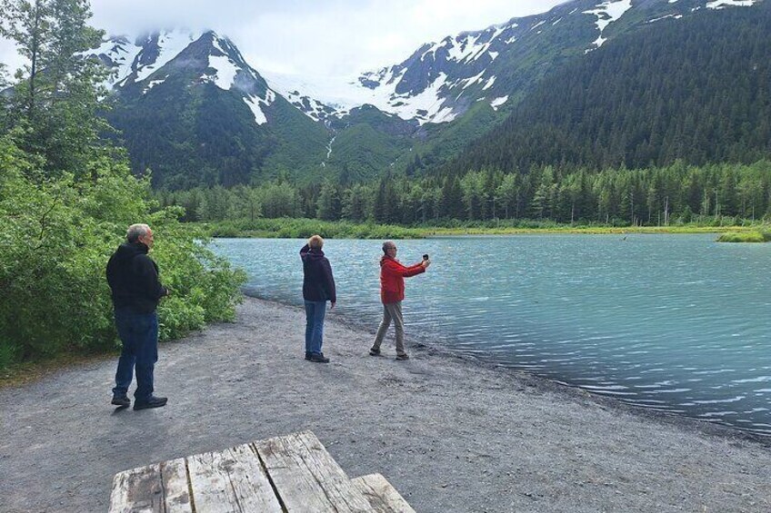 Drei Personen stehen an einem See, im Hintergrund Berge. 1