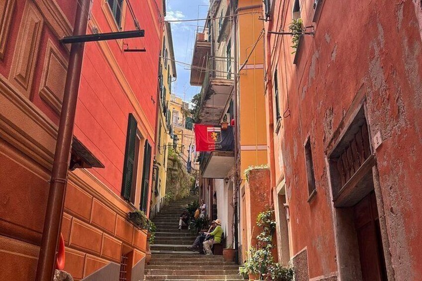 A narrow street with buildings on either side, a person descending stairs, and a red flag hanging from a building. 2