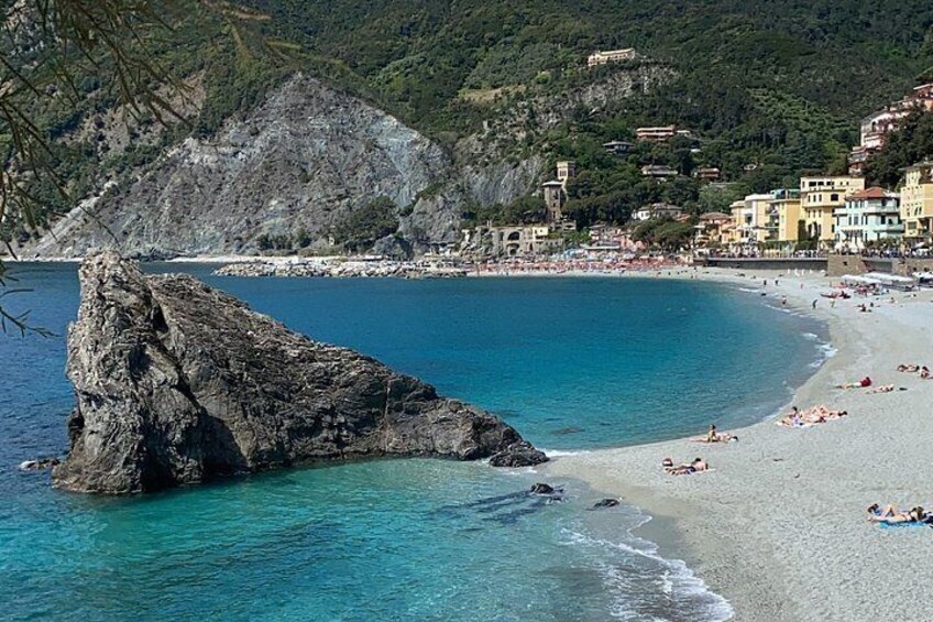 A coastal beach with clear blue waters, a prominent rock formation, and buildings along the shore. 4