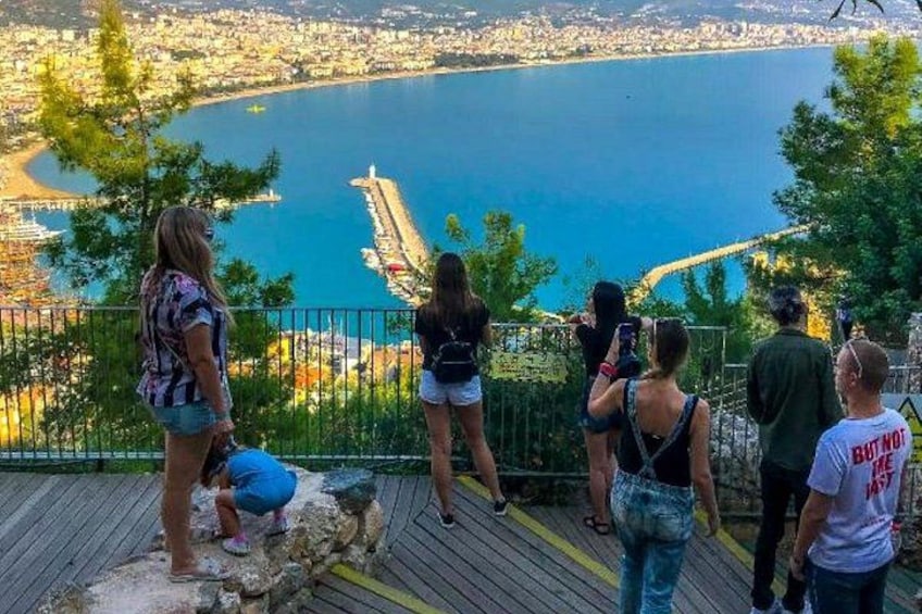 A group of people on a wooden deck overlooking a coastal city with a clear view of the sea and a pier. 5