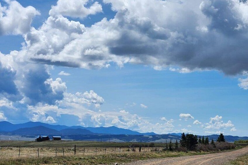 A rural landscape with a dirt road, a fence, and distant mountains under a partly cloudy sky. 4