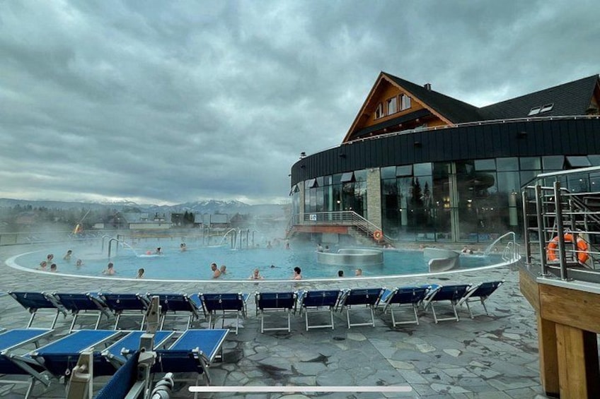 A hot spring pool with people relaxing, a wooden building, and a cloudy sky. 4