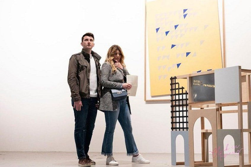 Two people standing in an art gallery with a large yellow poster and a wooden structure with a grid pattern. 5