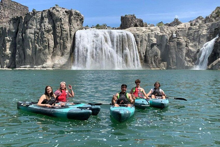Menschen paddeln mit ihren Kajaks auf einem ruhigen Fluss, im Hintergrund ein Wasserfall. 4