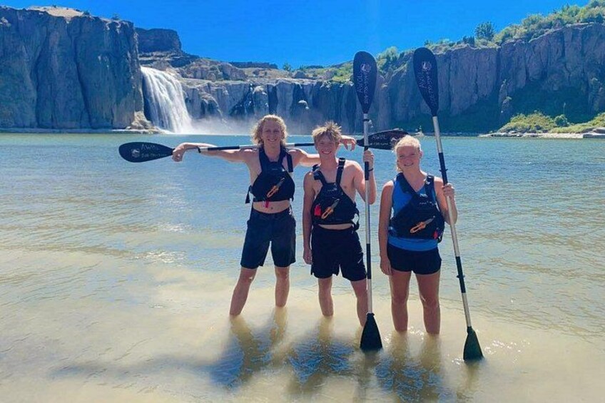 Drei Personen stehen in flachem Wasser mit Kajaks, ein Wasserfall ist im Hintergrund zu sehen und der Himmel ist wolkenlos. 5