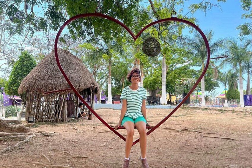 A person standing in a heart-shaped frame with a thatched hut and palm trees in the background. 2
