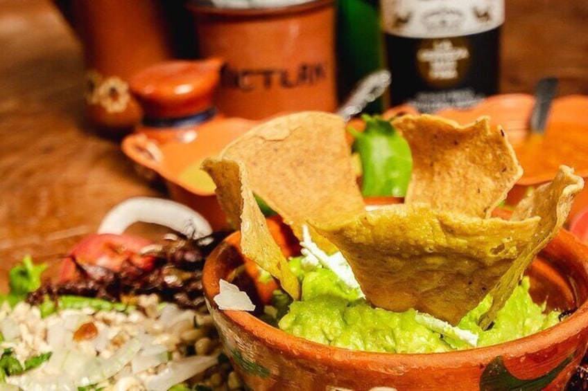 A bowl of guacamole with a tortilla chip on top, a side salad, and a bottle of sauce in the background. 3