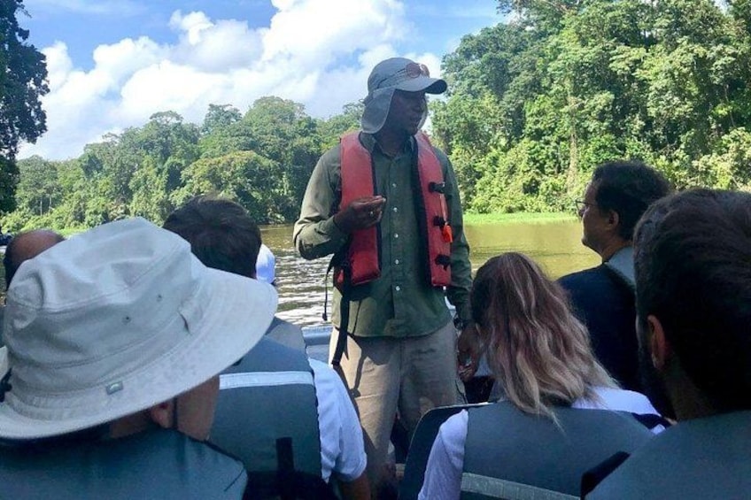 A group of people on a boat, a guide in a red vest, and a forested landscape in the background. 1