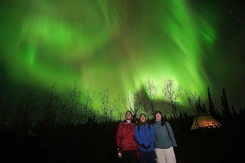 Three people standing under the aurora borealis with trees and a tent in the background. 4