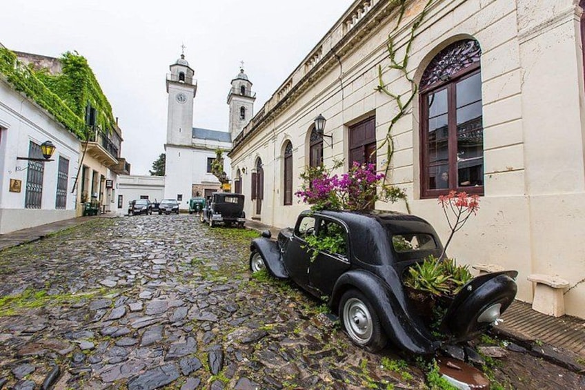 Una calle adoquinada con un coche antiguo estacionado a un lado, una iglesia con dos torres y edificios cubiertos de hiedra y flores. 1