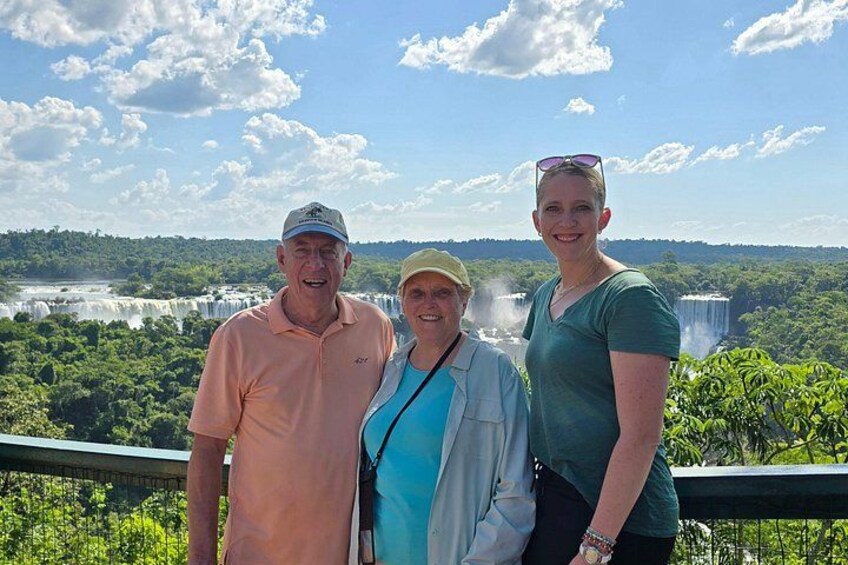 Three people standing on a bridge with a waterfall and greenery in the background. 2