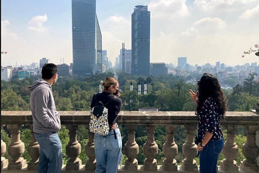 Three people standing on a balcony overlooking a cityscape with tall buildings and greenery. 1