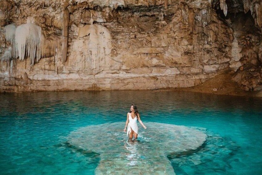 Eine Person in einem weißen Kleid steht in einem klaren, blauen Pool, umgeben von steinernen Felswänden. 1