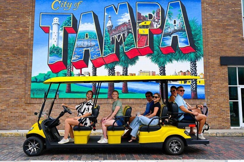 A yellow golf cart with passengers, a mural of Tampa, and a brick building in the background. 1