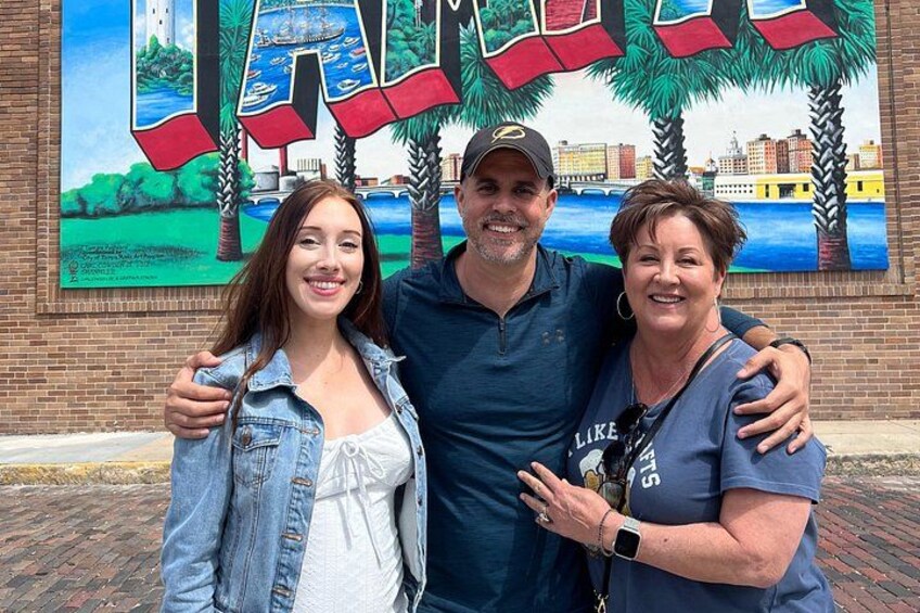 Three people standing in front of a mural with palm trees and water. 4