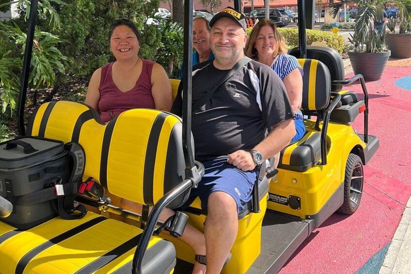 A yellow golf cart with passengers, a black bag, and a red and black striped bag. 2
