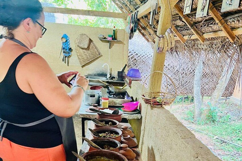 A person cooking outdoors with pots and pans on a table, surrounded by a rustic setting with a thatched roof and wooden structures. 5