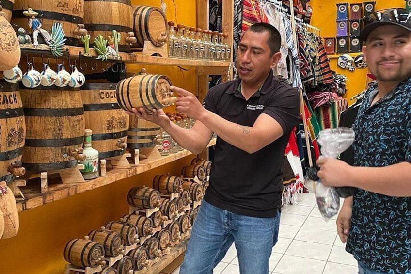 Eine Person hält eine Spirituosenflasche, im Hintergrund hölzerne Fässer und verschiedene Flaschen auf Regalen in einem Laden. 4