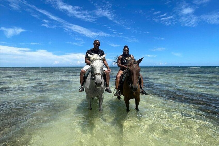 Two people riding horses in shallow water with a clear sky. 4