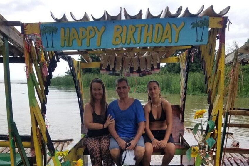 A birthday celebration setup with a 'HAPPY BIRTHDAY' sign, a wooden structure, and a body of water in the background. 2