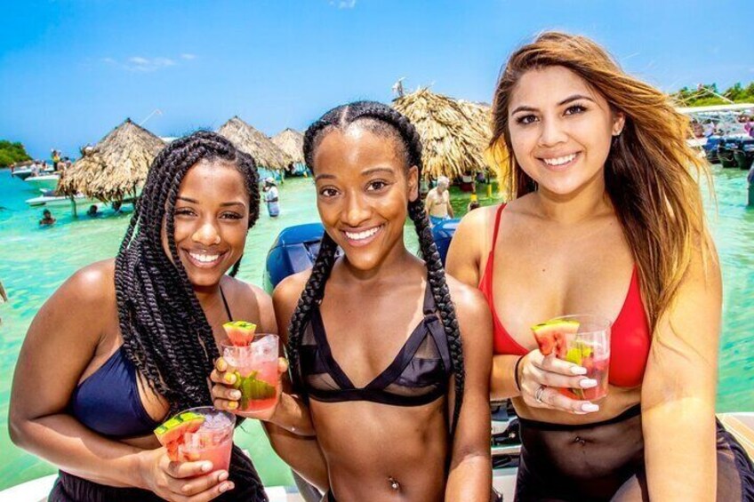 Three people on a boat with drinks, tropical beach, and thatched umbrellas in the background. 1