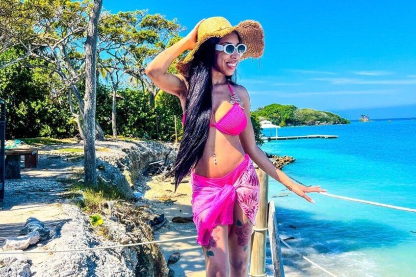 A person in a pink bikini and straw hat by a clear blue sea with rocky terrain and trees. 3