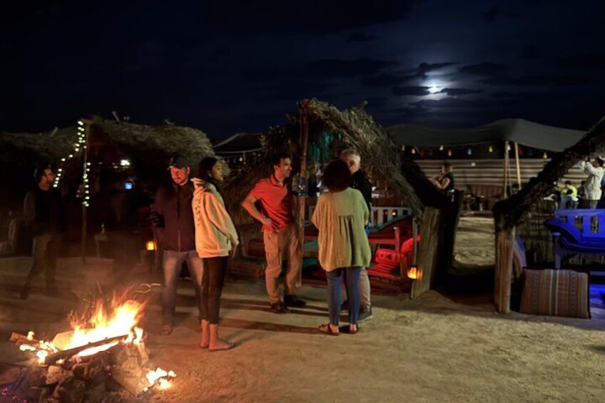 A group of people gathered around a campfire at night, with a thatched-roof structure and string lights in the background. 5