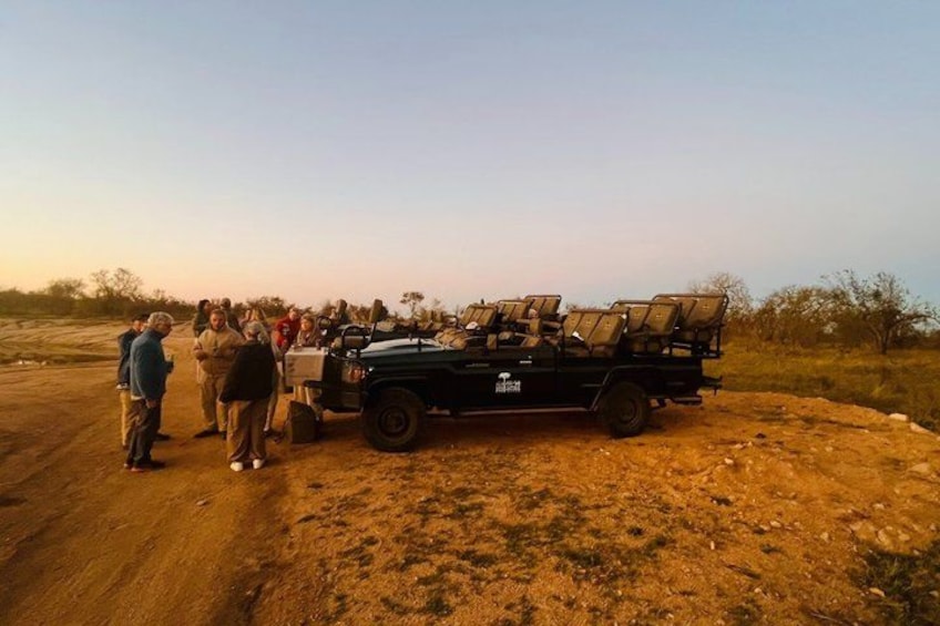 A group of people gathered around a black off-road vehicle with a roof rack, parked on a dirt road in a dry, open landscape with sparse vegetation. 2