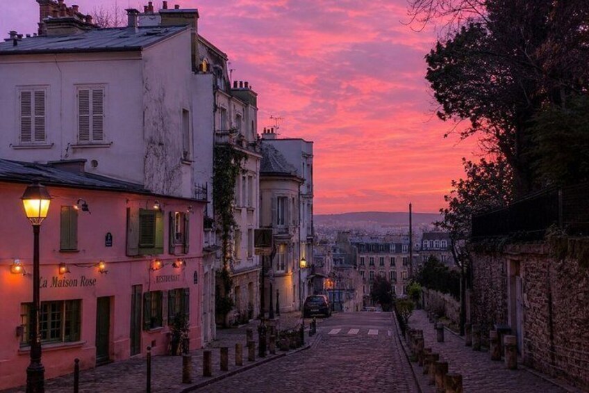 A cobblestone street lined with buildings, a pink building with 'La Maison Rose' signage, and a sunset sky. 2
