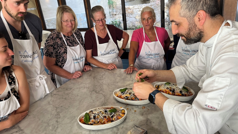 Ryhmä ihmisiä, joilla on yllään esiliinat, joissa on painettuna teksti »Towns of Italy Cooking School», valmistaa ruokaa marmorisella työtasolla. 1