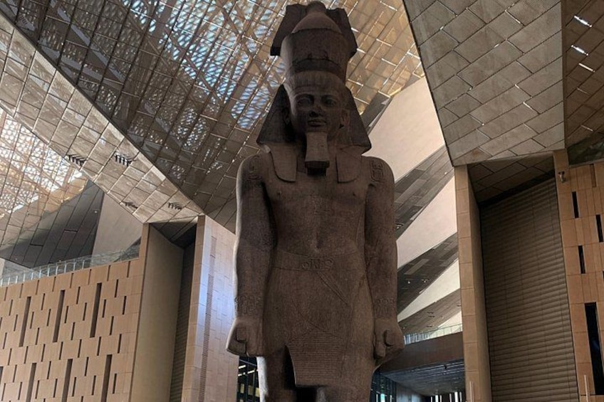 A large ancient Egyptian statue in a modern building with a patterned ceiling and wooden panel walls. 3