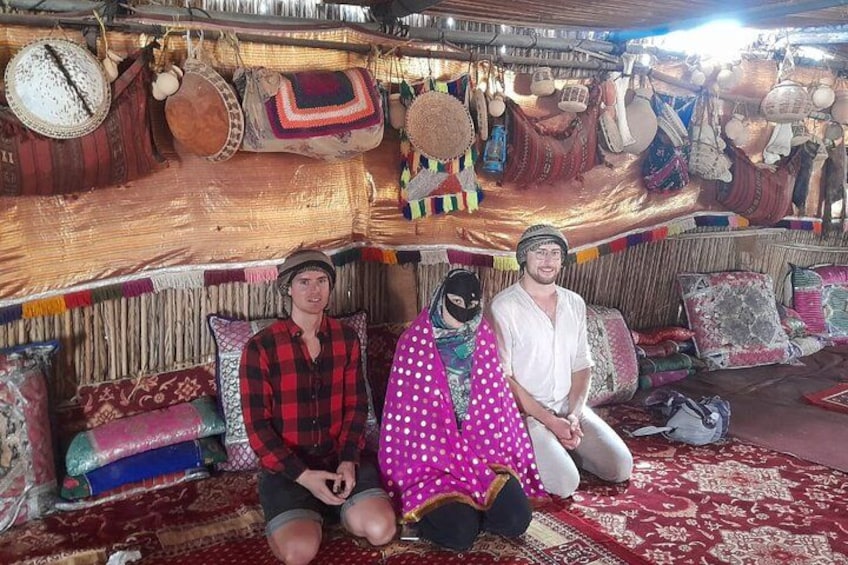 Two people sitting on a patterned rug in a tent-like structure with various textiles and bags hanging above them. 2