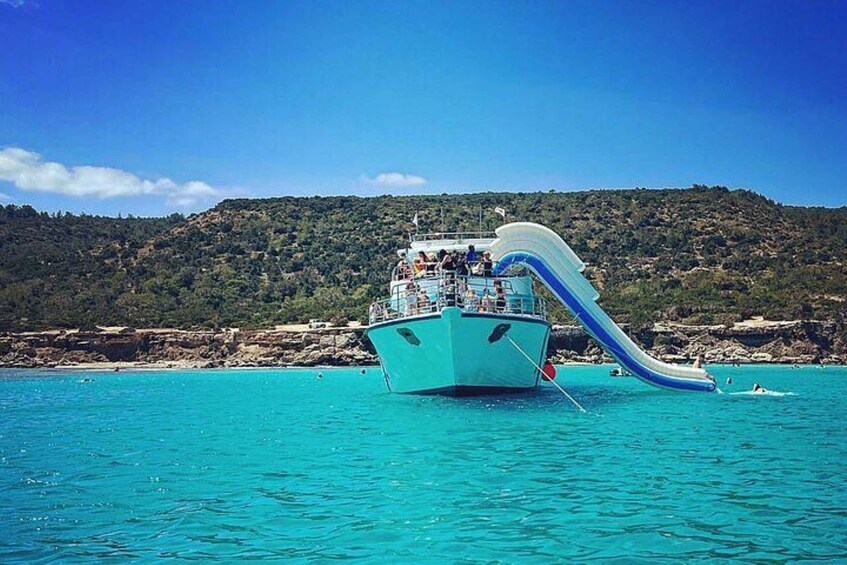 Un bateau doté d’un toboggan aquatique bleu, naviguant dans des eaux turquoise, entouré d’une côte rocheuse et de collines verdoyantes. 1