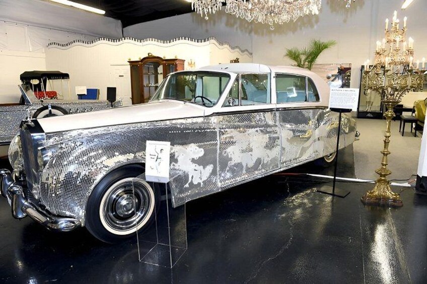A vintage car with a metallic finish, displayed indoors with a chandelier and a sign. 2