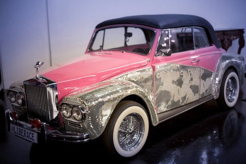 A vintage car with a pink and silver camouflage paint job, chrome details, and a 'LIBERACE' license plate. 2