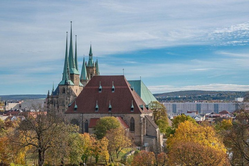 A historic church with multiple spires, surrounded by autumn-colored trees and a cityscape in the background. 2
