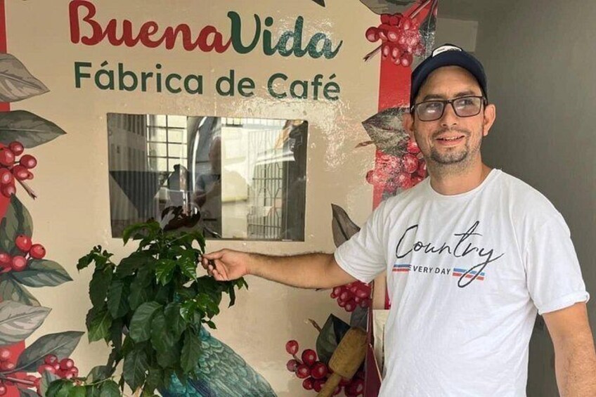 Une personne tenant une plante devant un mur sur lequel on peut lire le texte : « BuenaVida Fábrica de Café ». 2