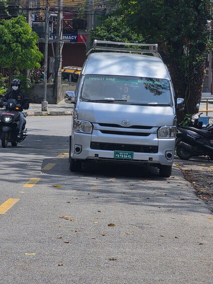A silver van with a 'TOURIST ONLY' sign on the windshield, parked on a street with yellow lines, near a tree and a pharmacy. 5