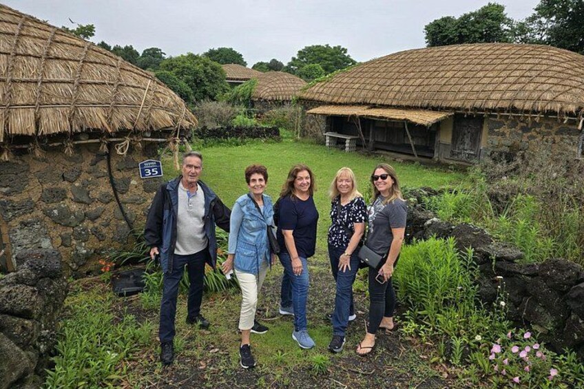A group of people standing in front of traditional thatched-roof huts with stone walls, surrounded by greenery and a small stream. 2