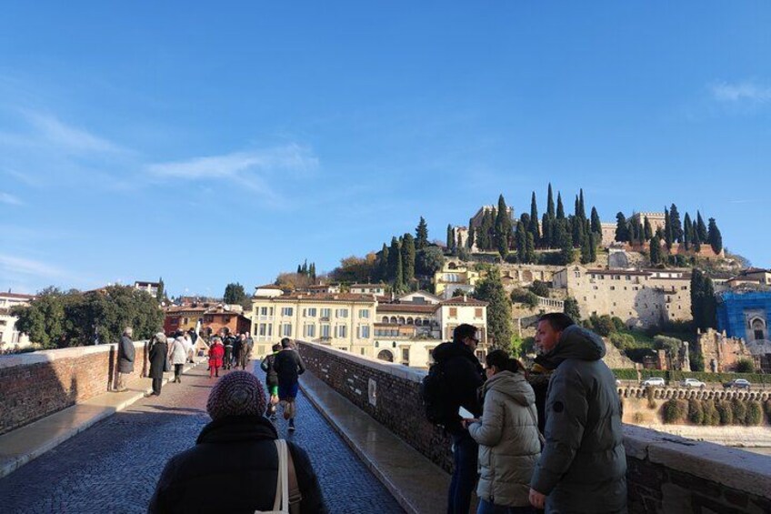 Un puente con gente caminando, un edificio histórico y una colina con árboles y una estructura parecida a un castillo al fondo. 4